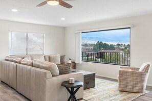 A modern living room with a beige sectional sofa, a small side table, a wicker chair, and a large window offering a view of a mountainous landscape. - Cottonwood Retreat ~ Large Swim Spa & Views, Games, Sauna (Cottonwood)