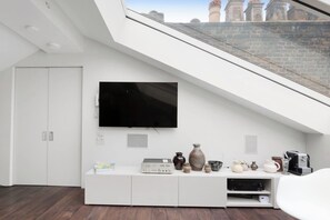 A modern living room with a flat-screen TV mounted on the wall, a white entertainment unit with various items, and a view of a brick wall and chimneys through a skylight.