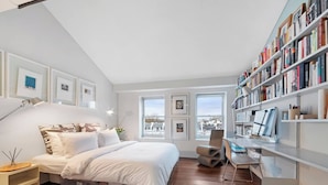 Een slaapkamer met een bed, nachtkastjes, een bureau met een computer, boekenplanken en ingelijste foto's aan de muur.