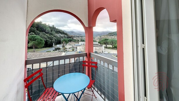 A balcony with a blue table and red chairs, overlooking a town with trees and hills.