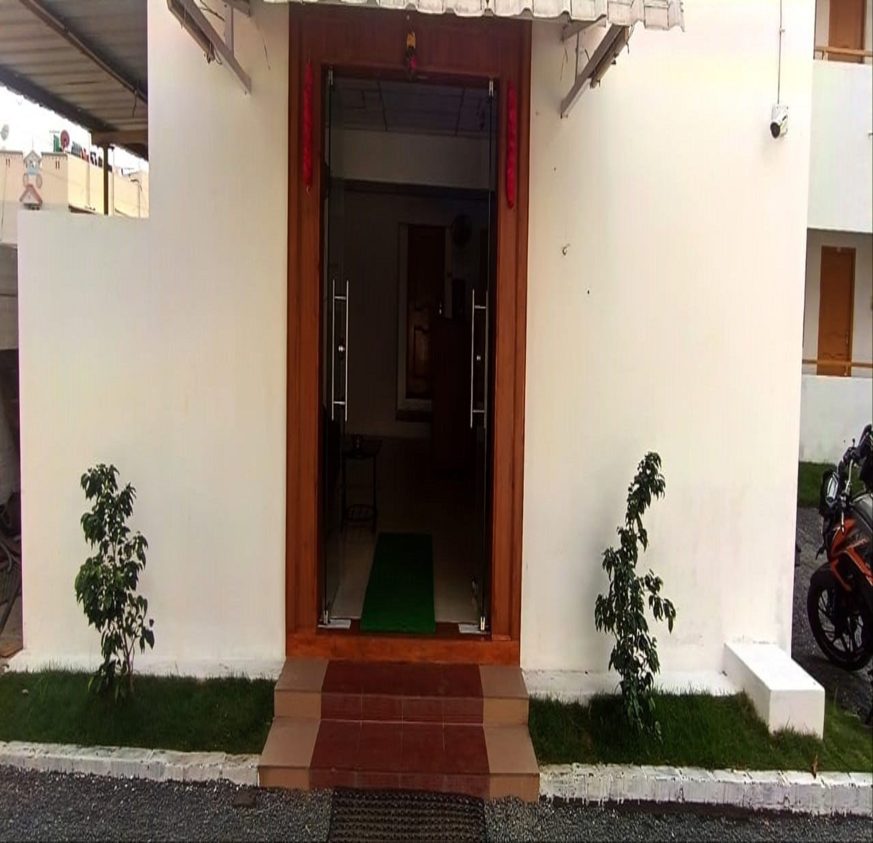 Une entrée d’immeuble avec une porte en bois, un paillasson vert et deux petites plantes de chaque côté.