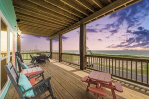 A wooden deck with picnic tables and chairs, overlooking a beach with a view of the ocean and a cloudy sky.