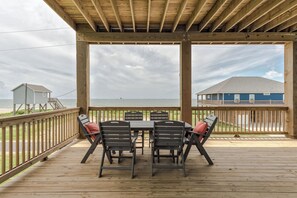 A covered outdoor deck with a table and chairs, overlooking a beach and a house.