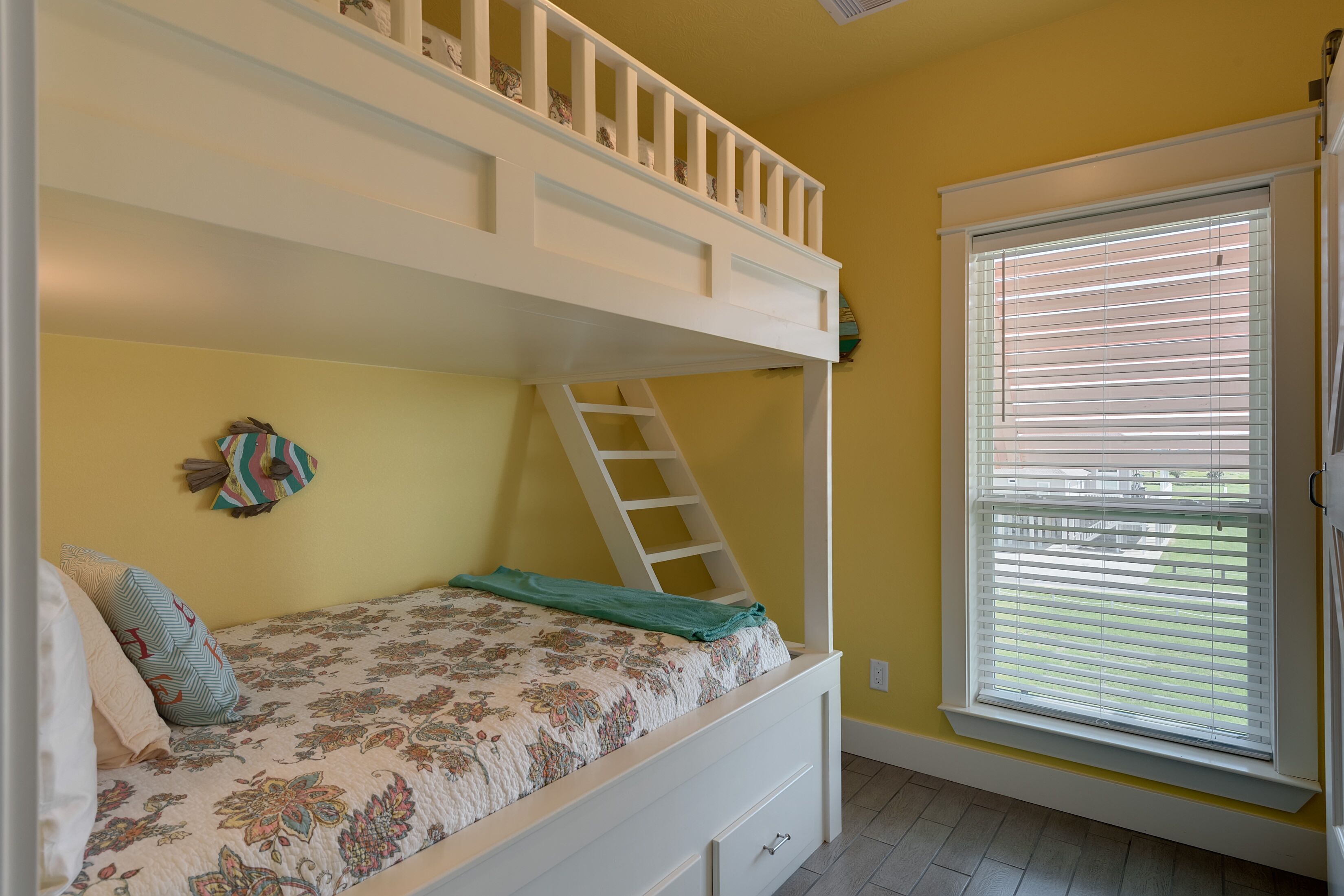Una cama litera con escalera, una ventana con persianas y una cama con colcha estampada.
