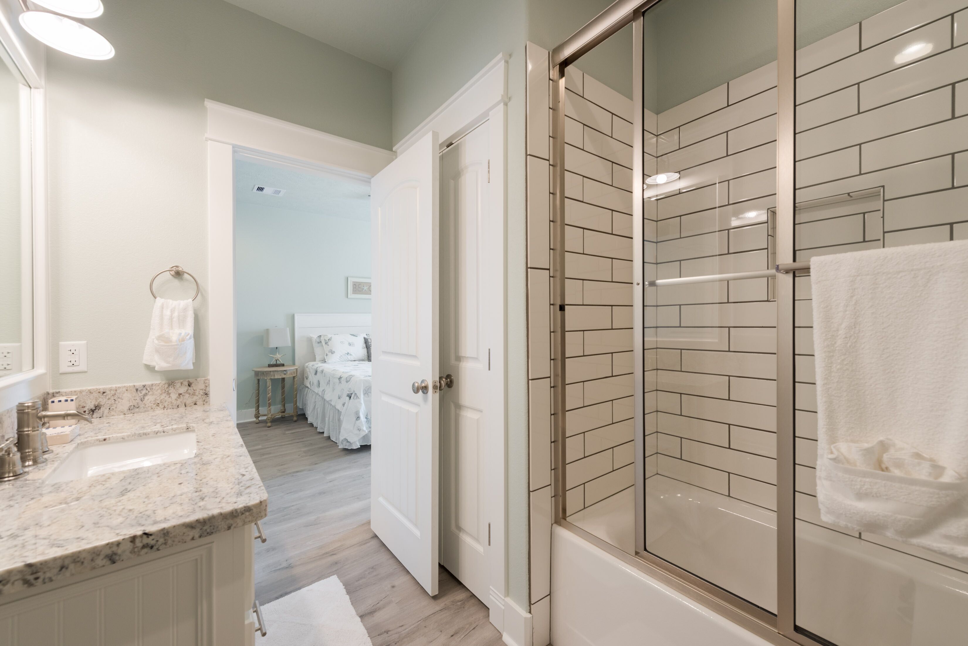 Un bagno con box doccia in vetro, un lavandino con piano in marmo e una porta bianca che dà su una camera da letto.