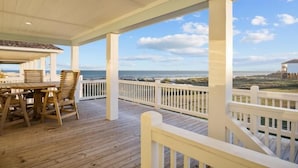 A covered outdoor deck with wooden furniture and white railings, overlooking a beach and ocean. - Gulf Views | Modern Amenities, Deck, Sleeps 12 & More (Port Bolivar)
