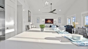 A modern dining area with a long table, a ceiling fan, and a fireplace in the background. - Gulf Views | Modern Amenities, Deck, Sleeps 12 & More (Port Bolivar)