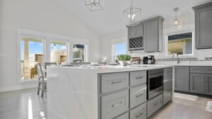 A modern kitchen with a large island, grey cabinets, and stainless steel appliances. - Gulf Views | Modern Amenities, Deck, Sleeps 12 & More (Port Bolivar)
