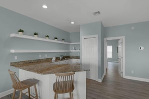 Una cocina con encimera de granito, armarios blancos y dos taburetes de madera. Hay estantes blancos con plantas en macetas, una puerta blanca y un pasillo que conduce a otra habitación.