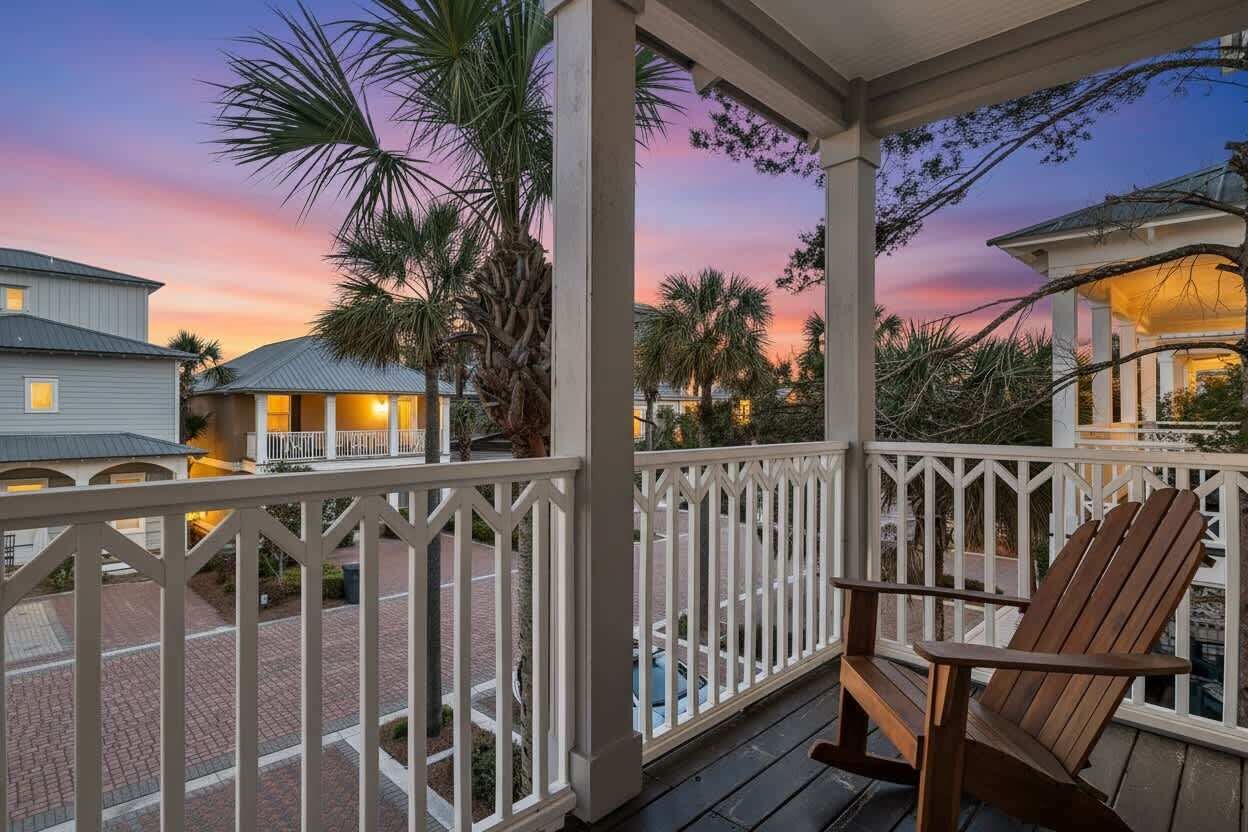 Eine Veranda mit Holzstuhl, weißer Balustrade und Blick auf Nachbarhäuser und Palmen bei Sonnenuntergang.