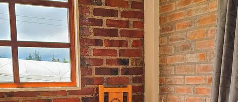 Una habitación con pared de ladrillo, ventana, cama con una manta de colores y una silla de madera.