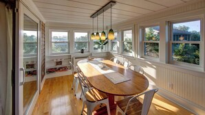 A dining area with a wooden table and chairs, large windows with views of greenery, and pendant lights. - Lucky Tern - Ocracoke Island (Ocracoke)
