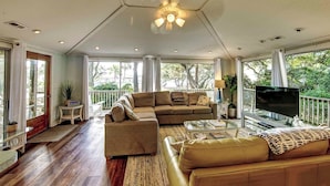 Un amplio salón con un sofá modular grande, una mesa de centro de vidrio y un televisor de pantalla plana. La habitación tiene un techo alto con un ventilador y luces empotradas.