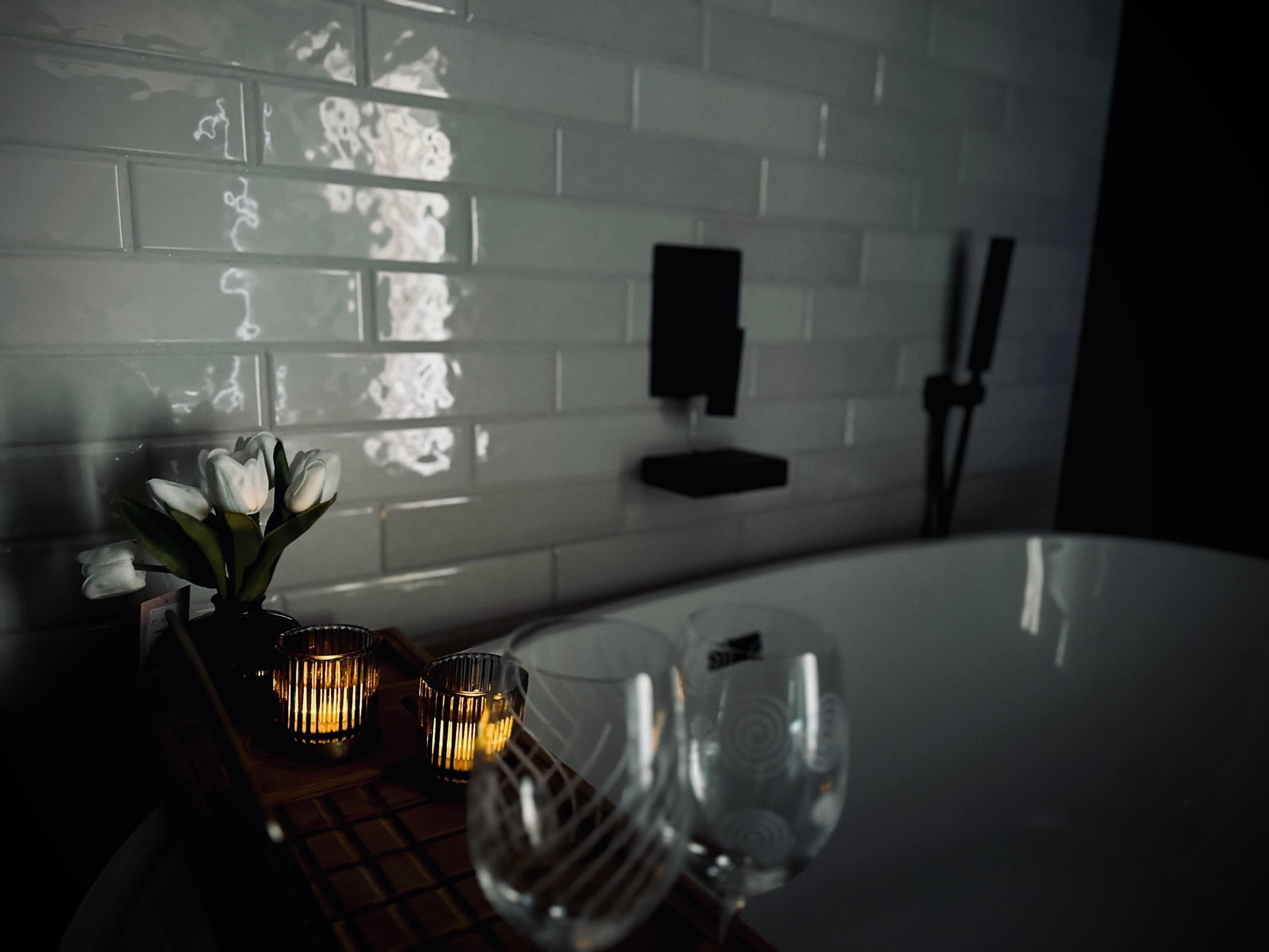 Une salle de bain avec un mur recouvert de carreaux blancs, une baignoire, un vase en verre contenant des fleurs blanches et deux bougies allumées.