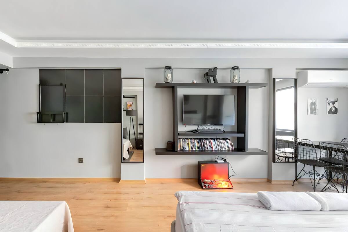 A modern hotel room with a large bed, a TV mounted on a wall, a bookshelf, and a dining area with chairs.