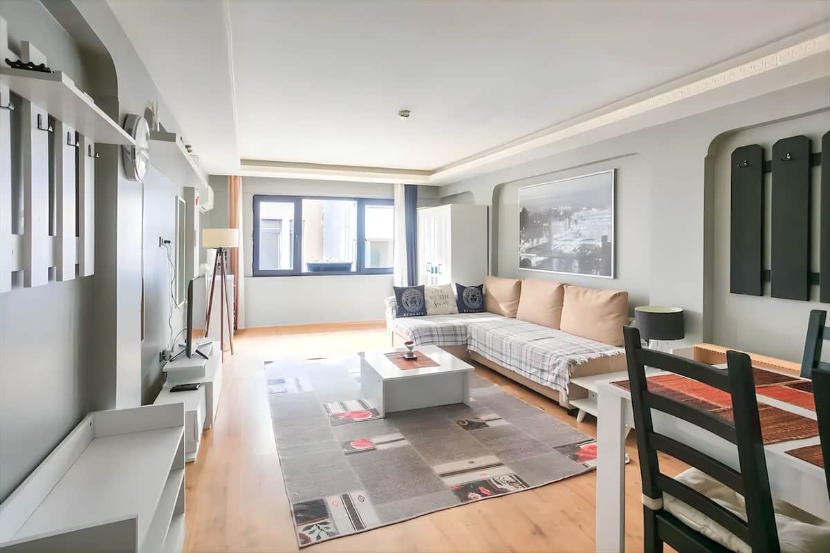 A modern living room with a sofa, coffee table, and a dining table with chairs. There is a large window providing natural light and a view of the city.