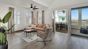 A spacious living room with a beige sofa, a grey armchair, and a coffee table. There is a large window with curtains, a ceiling fan, and a view of a balcony with a table and chairs. - Kas Ariba with pool and Laguna Marina View (Kralendijk)