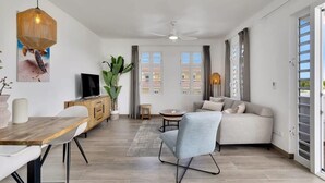 A modern living room with a sofa, dining table, and TV. There are windows with curtains, a ceiling fan, and a plant. - Kavel 17 - Turtles Place with a refreshing shared pool in the heart of Kralendijk (Kralendijk)