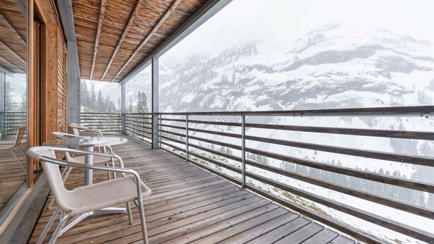 Une terrasse en bois, dotée de rampes et de chaises en métal, qui offre une vue sur un paysage montagneux enneigé.