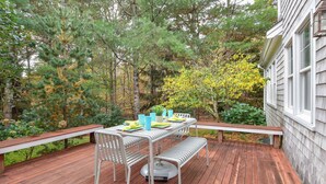An outdoor deck with a dining table set for four, surrounded by trees and a house.