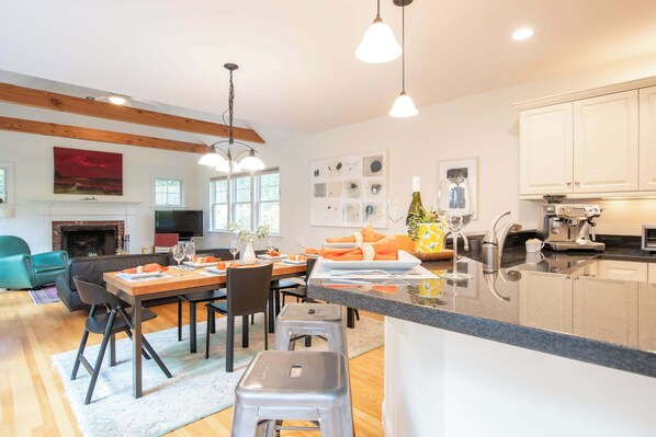 A modern kitchen with a central island, dining area with chairs and table, and a living room with a sofa and fireplace. - Perfectly private location w/deeded beach access! (Orleans)
