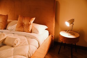 Una cama bien hecha con edredón blanco y almohadas, una mesita redonda con lámpara, y suelo de madera.