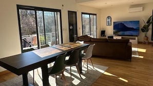 Ein geräumiges Wohnzimmer mit hoher Decke, großen Fenstern, einem Essbereich mit langem Tisch und Stühlen, einer Couch und einem Flachbildfernseher.