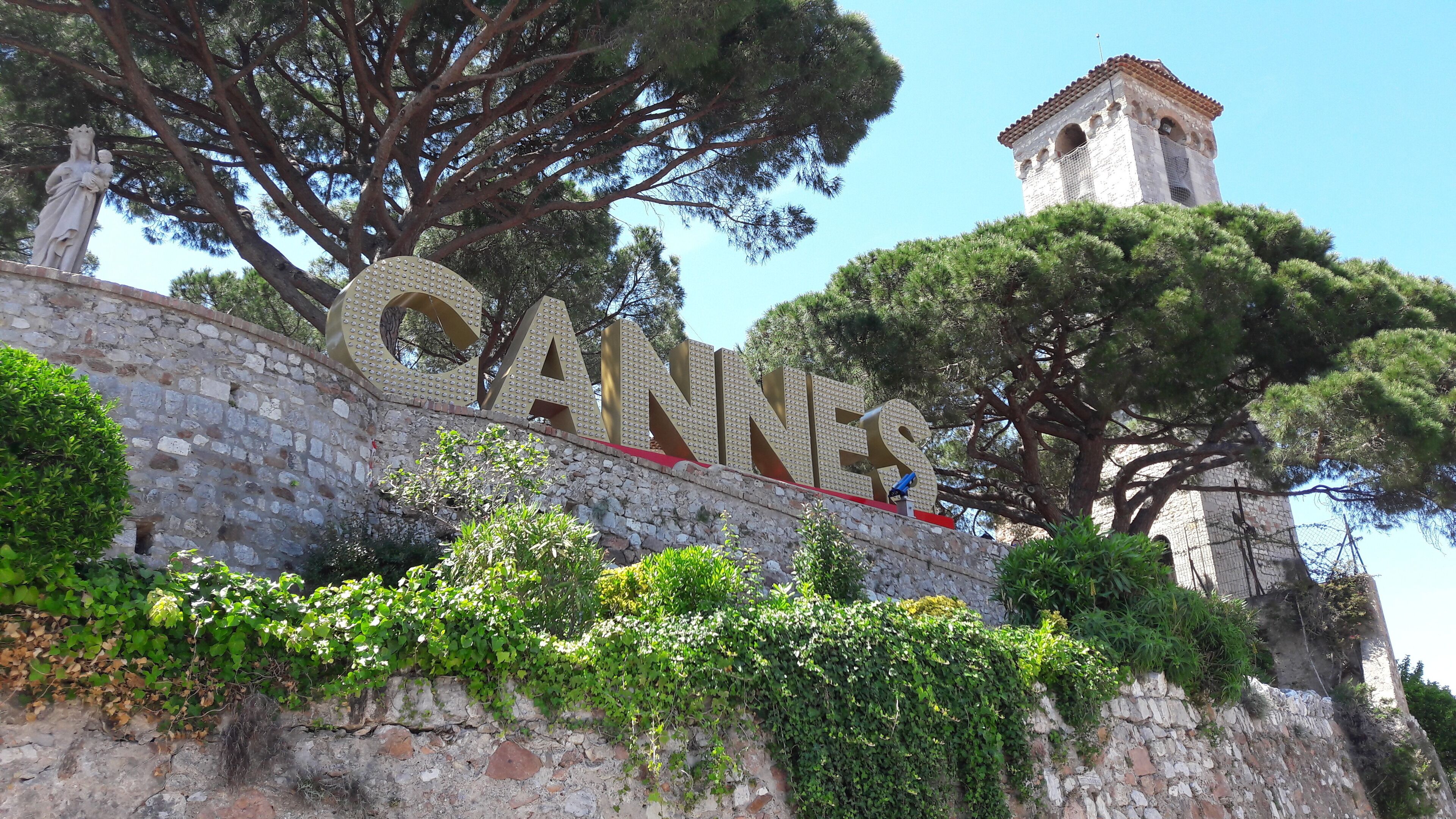 A stone wall with the word "CANNES" in large letters, a bell tower, and surrounding greenery.