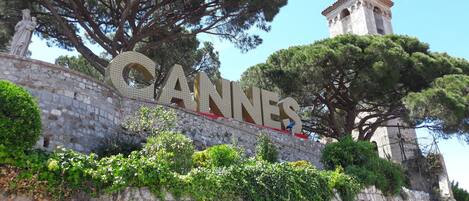 A stone wall with the word "CANNES" in large letters, a bell tower, and surrounding greenery.