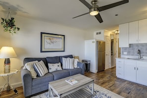 Un espace de vie compact comprenant un canapé gris, une table basse, un ventilateur de plafond, un coin cuisine avec des meubles blancs et un tableau encadré au mur.