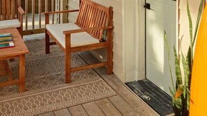 Una terraza de madera con un banco y una mesa, una puerta con buzón y una pequeña lámpara exterior.