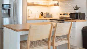A modern kitchen with white cabinetry, a wooden island with two chairs, a stainless steel refrigerator, a microwave, and a built-in oven.