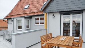 Une maison moderne avec une façade grise, un toit recouvert de tuiles rouges et une table et des chaises en bois sur le balcon.