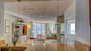 A kitchen with granite countertops, a stainless steel sink, pendant lights, and a dining area with a table and chairs. - Oceanfront Condo with Pool View (North Topsail Beach)