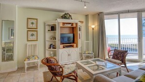 A living room with a television, wicker chairs, a glass coffee table, and a view of the beach through large windows. - Oceanfront Condo with Pool View (North Topsail Beach)