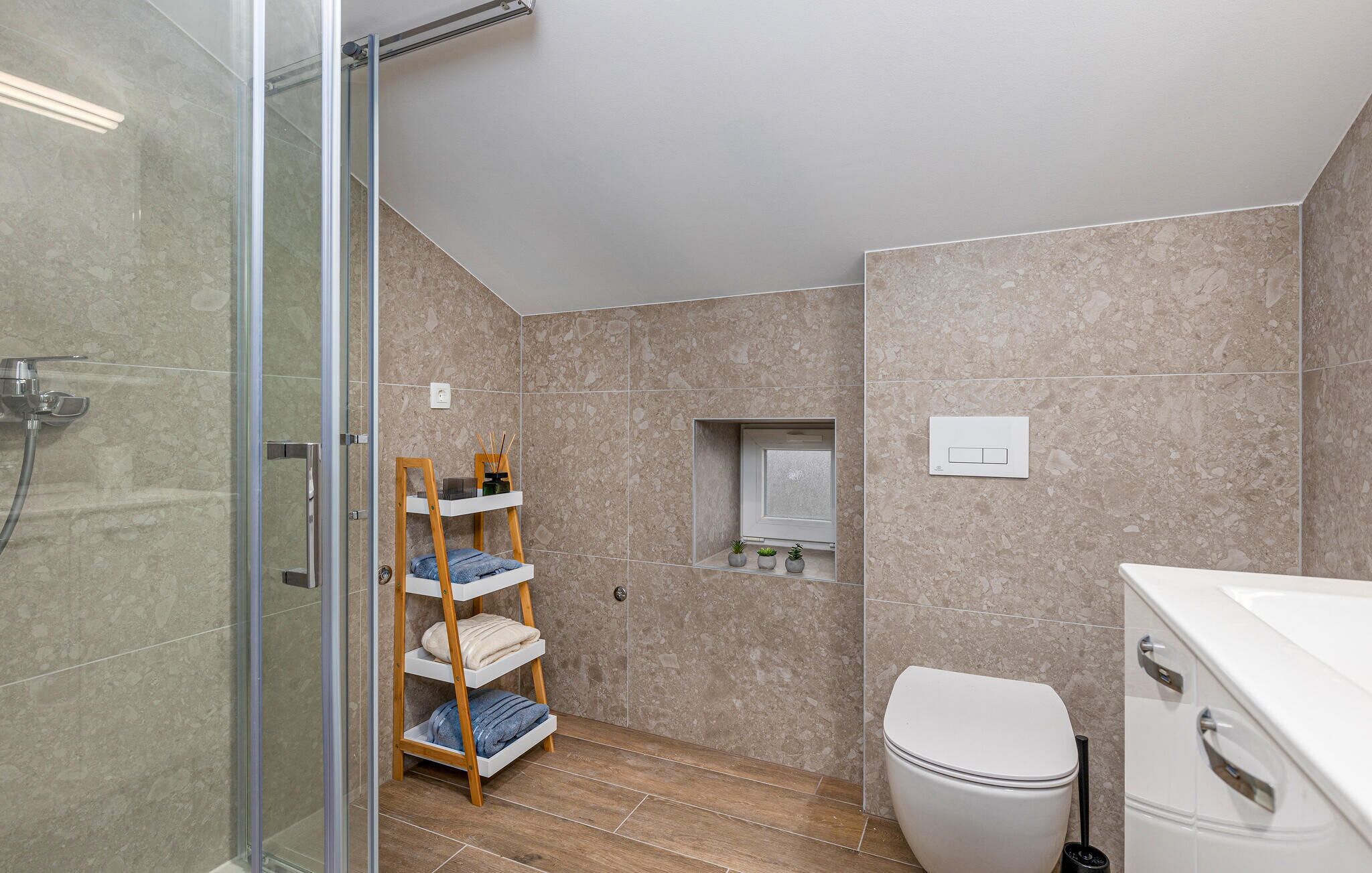 Une salle de bain moderne avec une douche à parois de verre, une étagère en bois où sont posées des serviettes, des toilettes et une petite étagère pour les plantes.