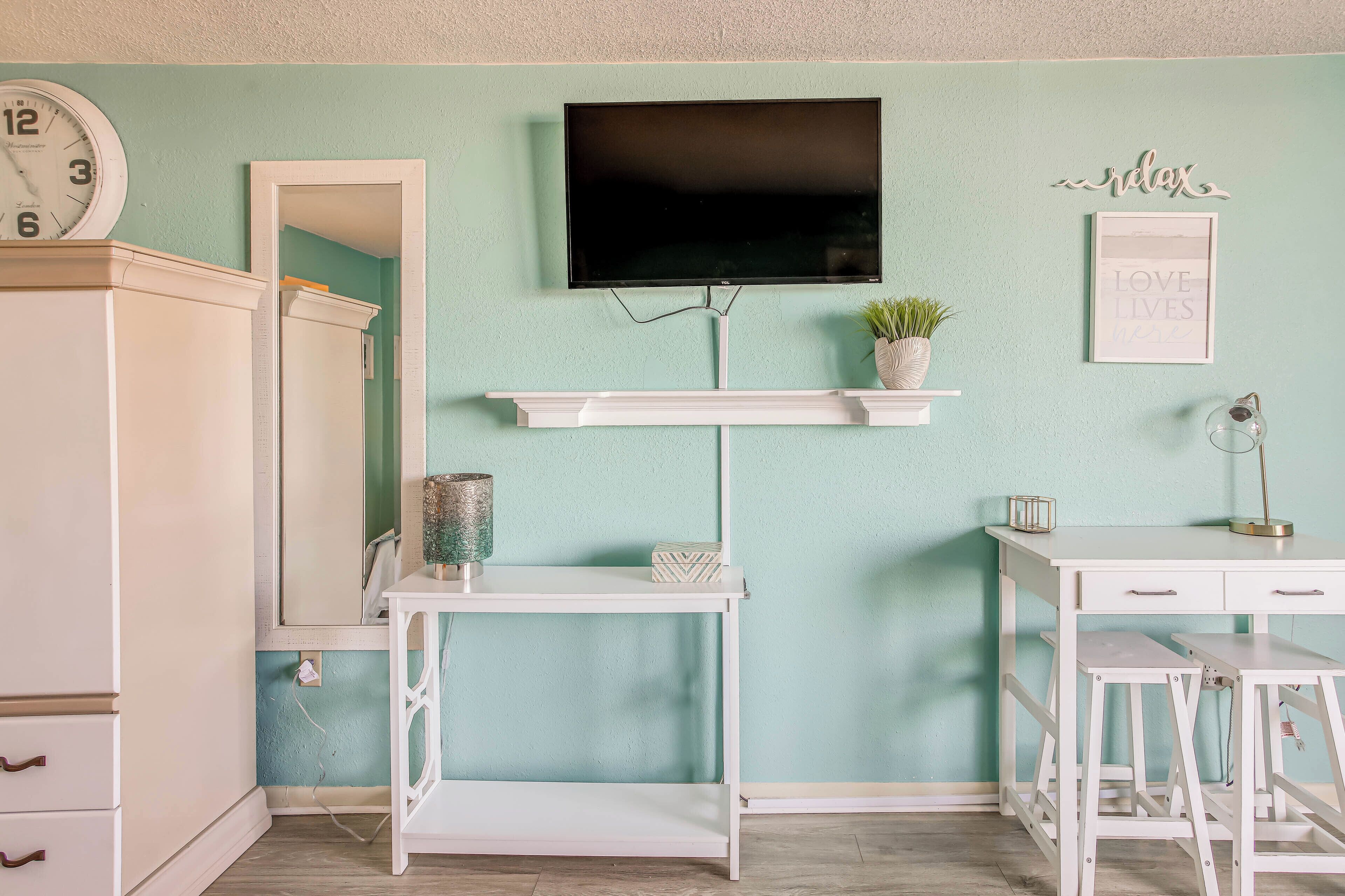 Una habitación con una pared color turquesa, un televisor de pantalla plana montado sobre una consola blanca, una planta pequeña en maceta y una cita inspiradora enmarcada.