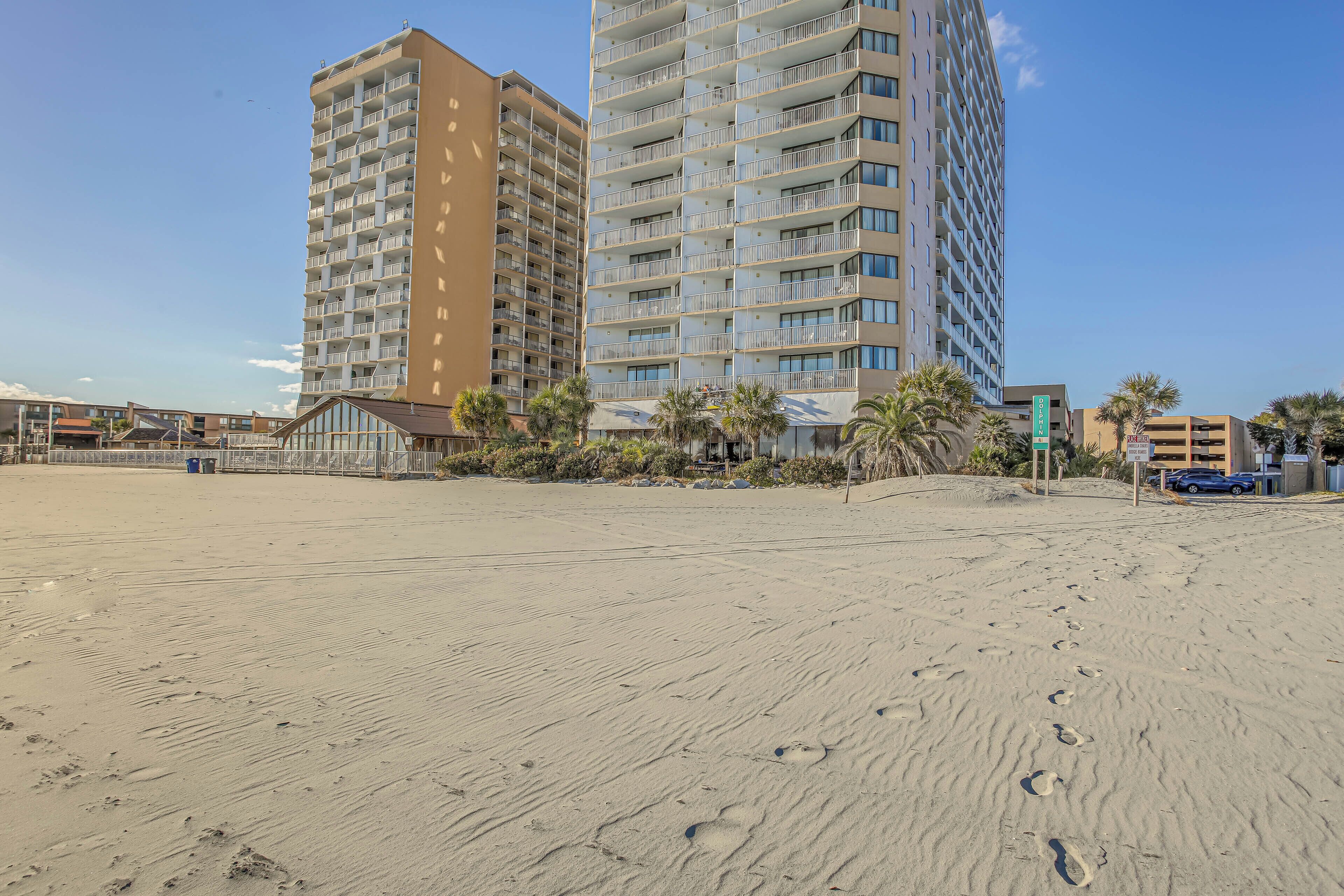 Una spiaggia con impronte che conducono a un edificio con diversi balconi e un cartello che indica un'area di parcheggio.