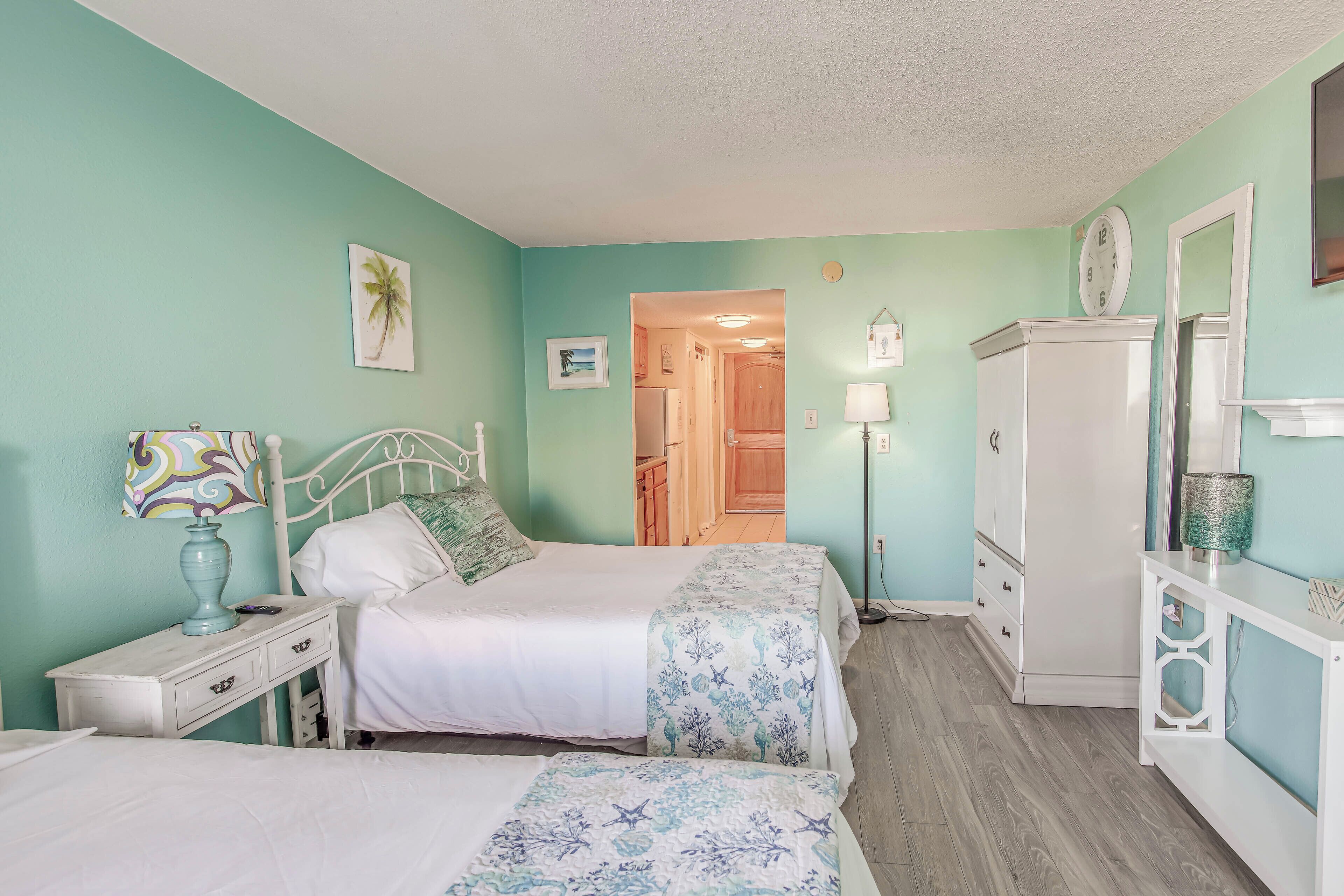 A bedroom with two beds, a nightstand, a dresser, and a door leading to a bathroom.