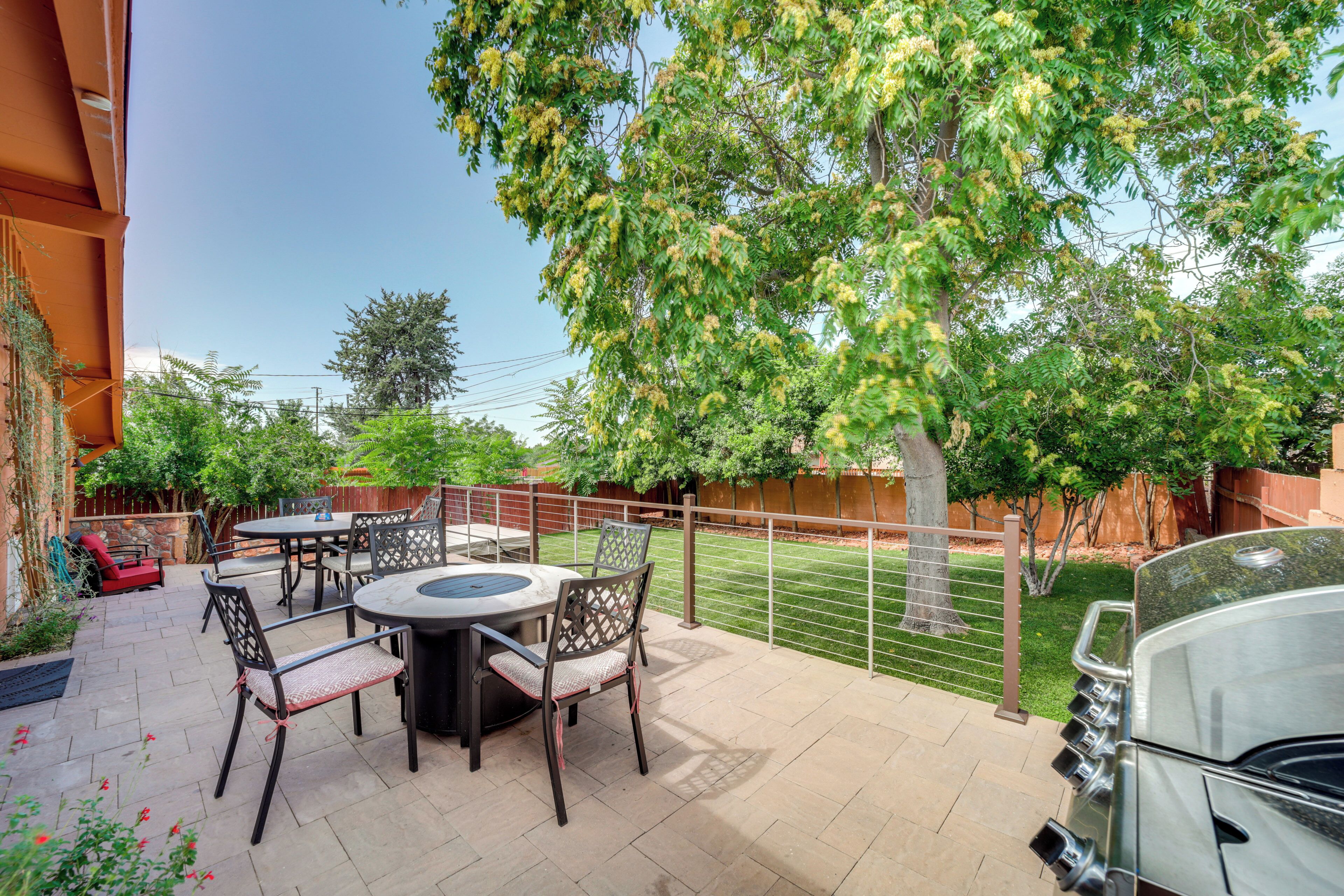 A patio with a round table and chairs, a barbecue grill, and a fenced yard with trees.