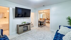 A modern living room with a flat-screen TV mounted on the wall, a blue sofa, a wooden entertainment unit, and a dining area in the background. - Michigan Retreat w/ Screened-in Porch, Near Lakes! (Belleville)