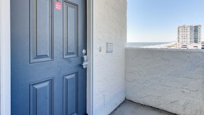 Eine blaue Tür mit Schlüsselloch und Schloss, eine weiße strukturierte Wand und im Hintergrund ein Strand sowie Gebäude.