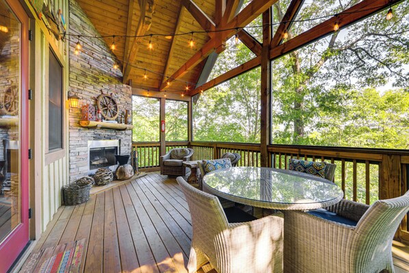 A covered outdoor deck with wicker furniture, a glass-top table, and a stone fireplace. - Hike & Boat: Cozy Bear Lake Cottage! (Tuckasegee)