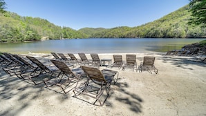 Ein Strand mit Reihen aus Metallstühlen und -tischen, umgeben von bewaldeten Hügeln und einem stillen See.