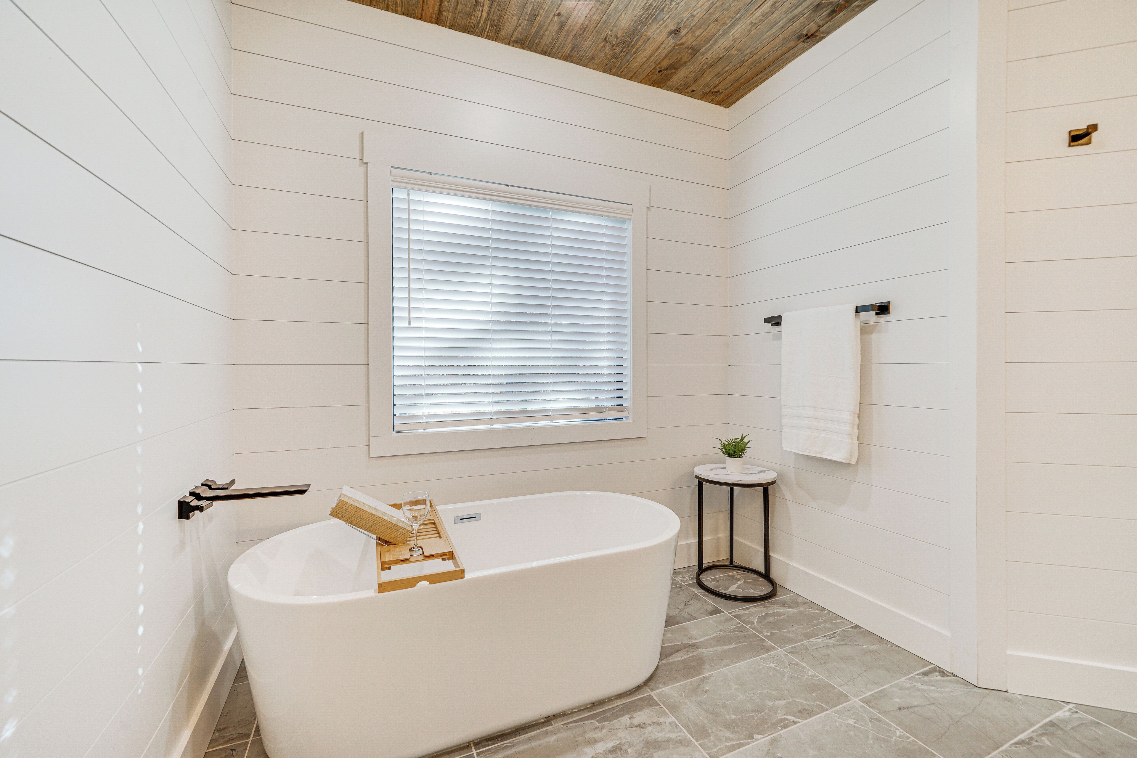 A modern bathroom with a freestanding bathtub, a small round table with a plant, and a window with blinds.