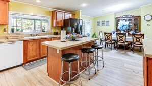Une cuisine avec des meubles en bois, un grand îlot central doté de tabourets, un coin repas avec des chaises et une horloge au mur.