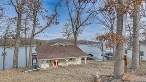 A house with a porch overlooking a lake, surrounded by trees and a gravel driveway. - Private Dock: Lake of the Ozarks Hideaway! (Sunrise Beach)