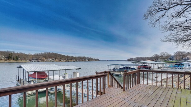A wooden deck with a railing overlooking a calm body of water, docks with boats, and a distant shoreline with buildings. - Private Dock: Lake of the Ozarks Hideaway! (Sunrise Beach)
