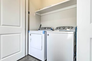 A laundry room with two white front-loading washers and dryers, a white door, and a white shelf above the dryers. - Inlet Beach Cottage: Close to Beach Access! (Panama City Beach)