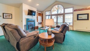 A spacious living room with two reclining leather sofas, a wooden coffee table, a fireplace, and a large window with a view of trees. - 17 Mi to Skiing! Lake-view Michigan Home w/ Deck (Harbor Springs)
